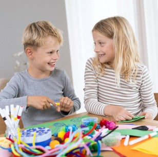 Two children sitting at a table with creative craft materials, smiling and engaged in an activity.