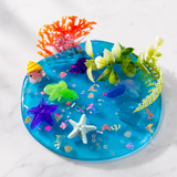 A round blue resin tray filled with colorful sea-themed decorations, including starfish, coral, small fish, and artificial plants, photographed on a light marble surface.