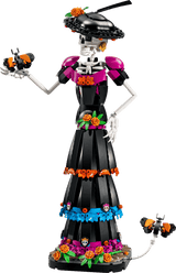 A Day of the Dead skeleton figurine built from LEGO, featuring a black dress with pink, orange, and blue floral accents, a black hat with flowers, and skull details, shown against a solid green background.