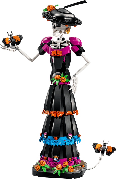 A Day of the Dead skeleton figurine built from LEGO, featuring a black dress with pink, orange, and blue floral accents, a black hat with flowers, and skull details, shown against a solid green background.