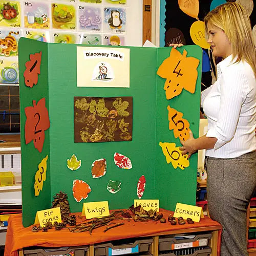 A woman stands beside a green educational display board labeled 'Discovery Table' in a classroom setting. The board features autumn-themed cutouts, numbered leaf shapes, a leaf collage, and labels for 'Fir cones,' 'twigs,' 'leaves,' and 'conkers' placed on a table below.