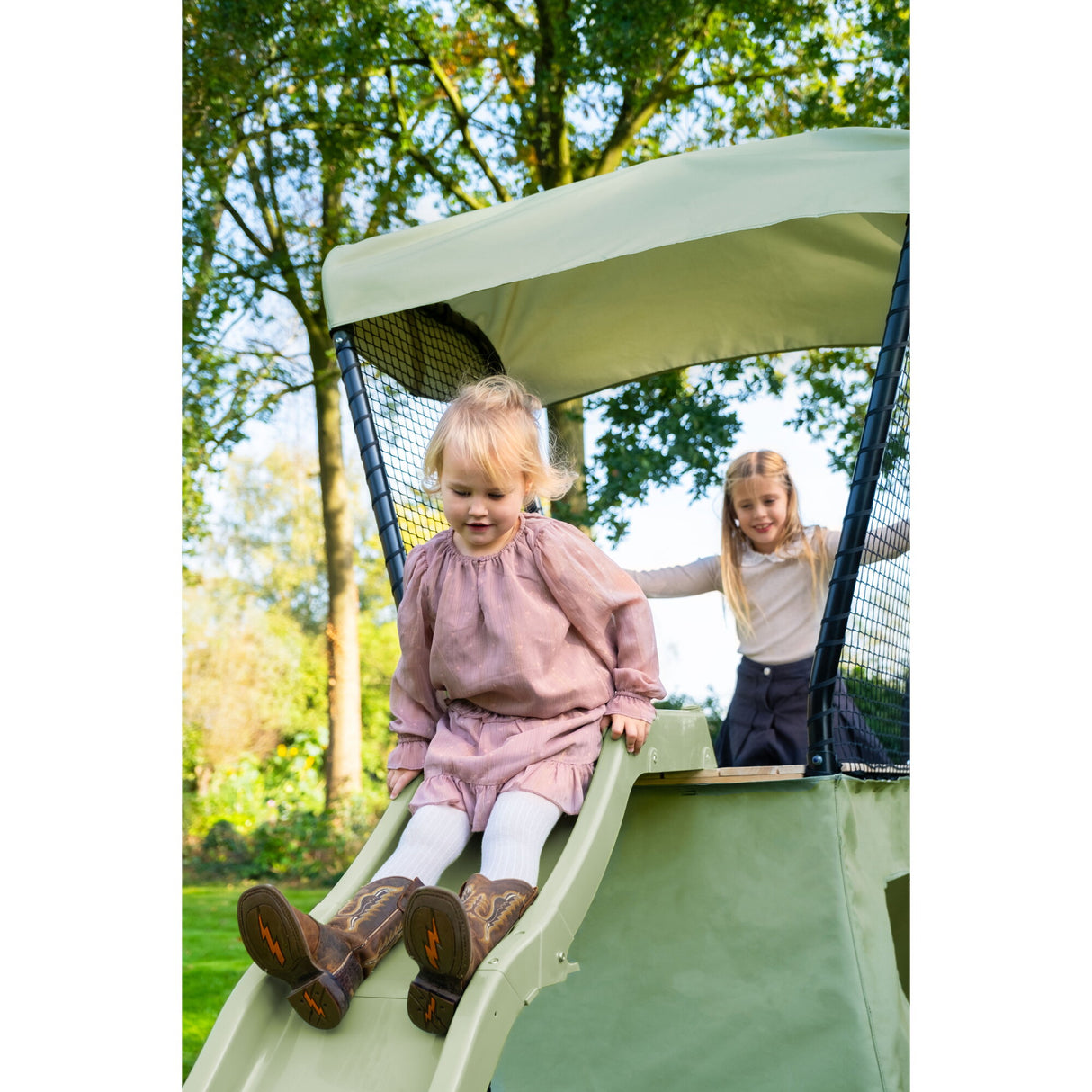 Two children sliding down a green slide with a canopy in an outdoor setting.