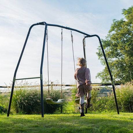 Person standing next to a swing set in a garden with trees and grass in the background