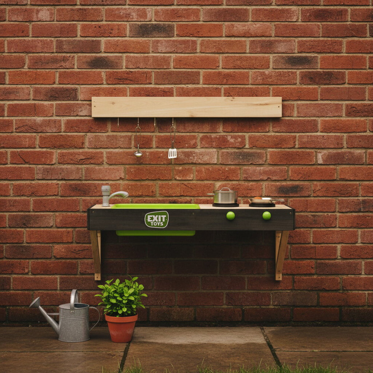Play kitchen set against a brick wall with a small plant and watering can on the ground.