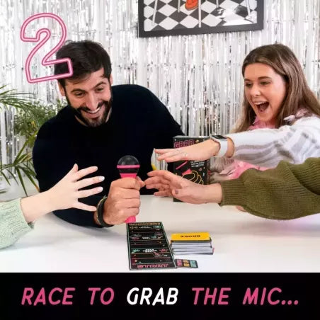 Two people playing a game with a microphone, surrounded by game cards on a table, with a festive background.