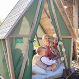 Woman sitting on a chair next to a wooden playhouse with two children inside, surrounded by firewood.