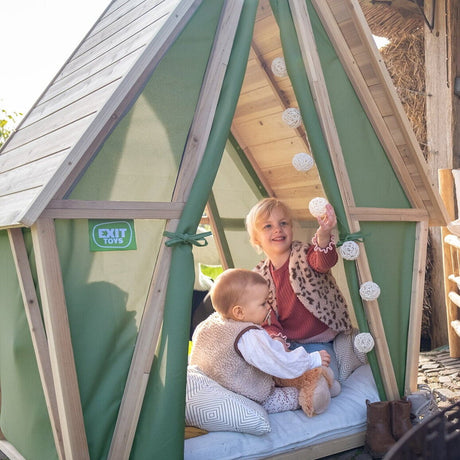 Woman sitting on a chair next to a wooden playhouse with two children inside, surrounded by firewood.